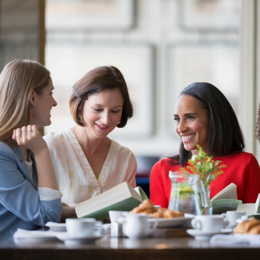Três mulheres conversando em torno de um livro aberto, em uma mesa de café